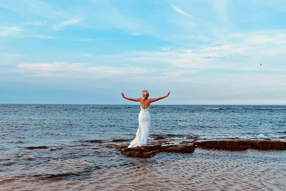 Bride stands on rocks at seaside, looking out to sea with arms outstretched